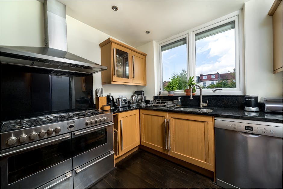 A modern kitchen featuring wooden cabinetry and black quartz countertops under natural light from two large windows. The stainless steel oven and stove are clean and polished, with a black backsplash
