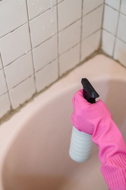 Close-up of a person wearing a bright pink rubber glove, holding a white spray bottle with a black nozzle, as they apply cleaning solution to the corner of a beige bathtub. The background features a t